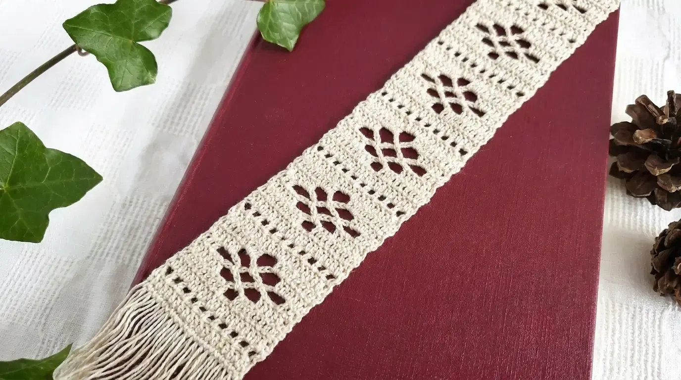 Ivory lace crochet bookmark with repeating openwork flowers and fringe laid diagonally across a burgundy book.